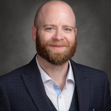 Man with a beard in a suit, smiling against a gray background.
