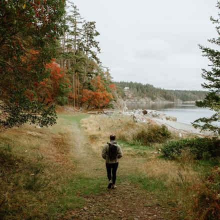 Person walking on a forest path by a river under an overcast sky.