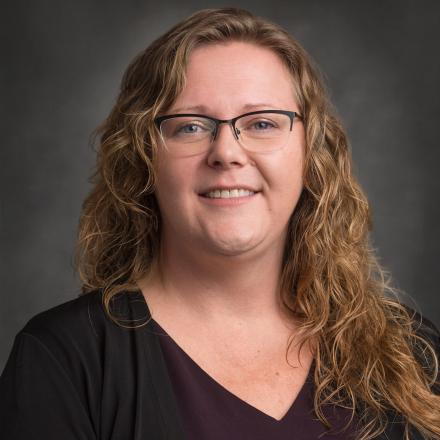 Smiling woman with glasses and wavy hair against a gray background.