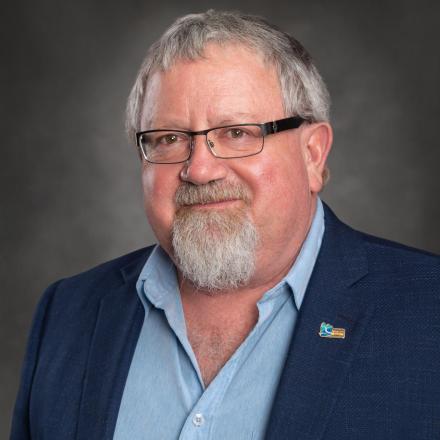 Bearded man in a suit and glasses, smiling against gray background.