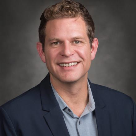 Smiling person in a suit jacket against a grey background.