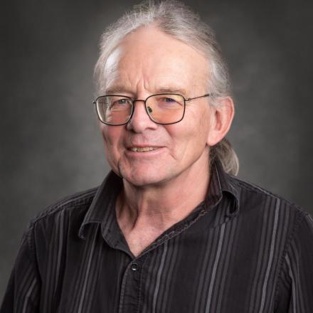Smiling man with glasses and long hair in a striped shirt, gray background.