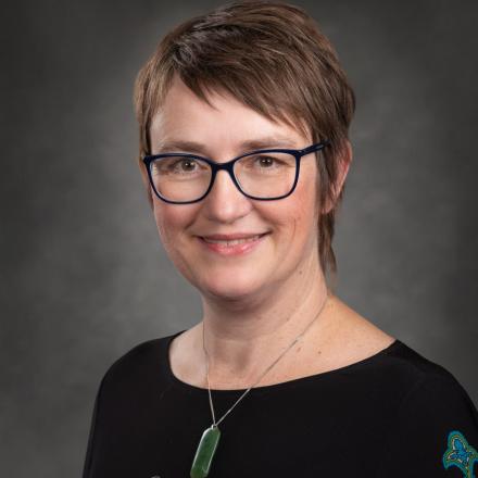 Smiling person with glasses, short hair, and a pendant, in a dark top with patterns.
