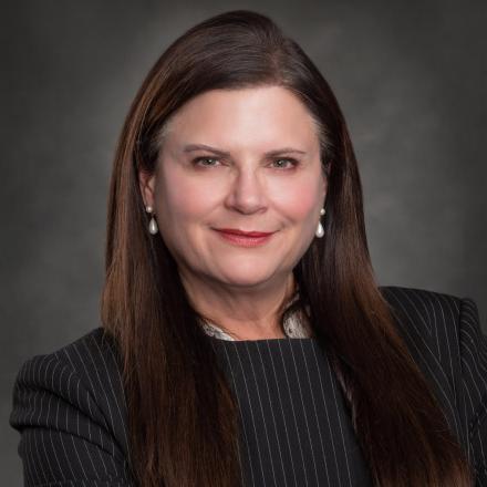 Dark-haired person in a pinstripe suit smiling against a grey background.