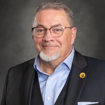 Smiling man in a suit with glasses and a goatee against a dark background.