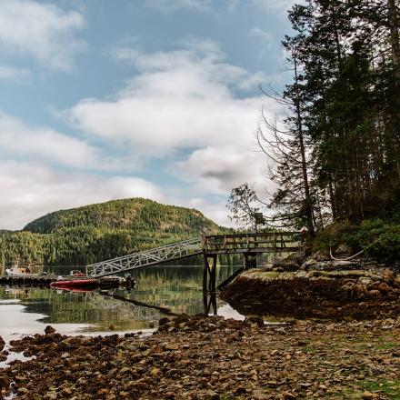 Calm lake with dock, surrounded by forest and hills under a cloudy sky.