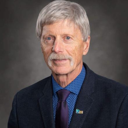 Ron Kerr | Older man with gray hair and mustache, wearing a suit against a gray background.