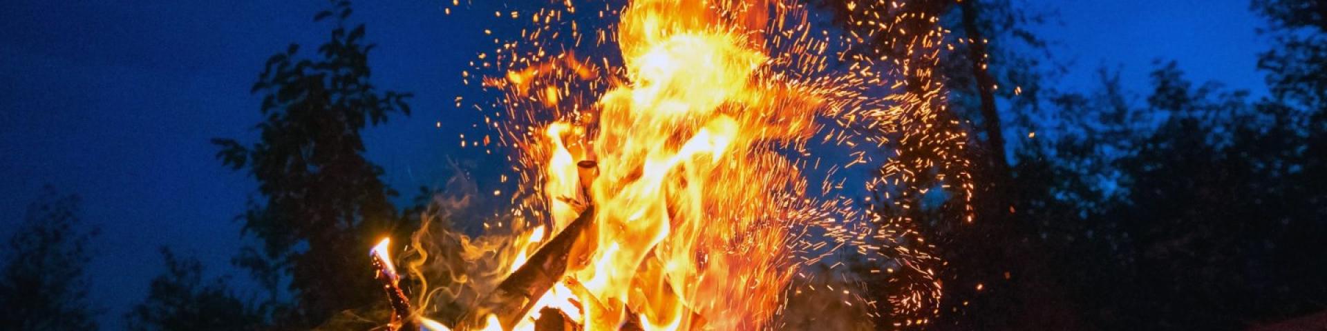 Campfire burning brightly at night, sparks rising against a deep blue sky.