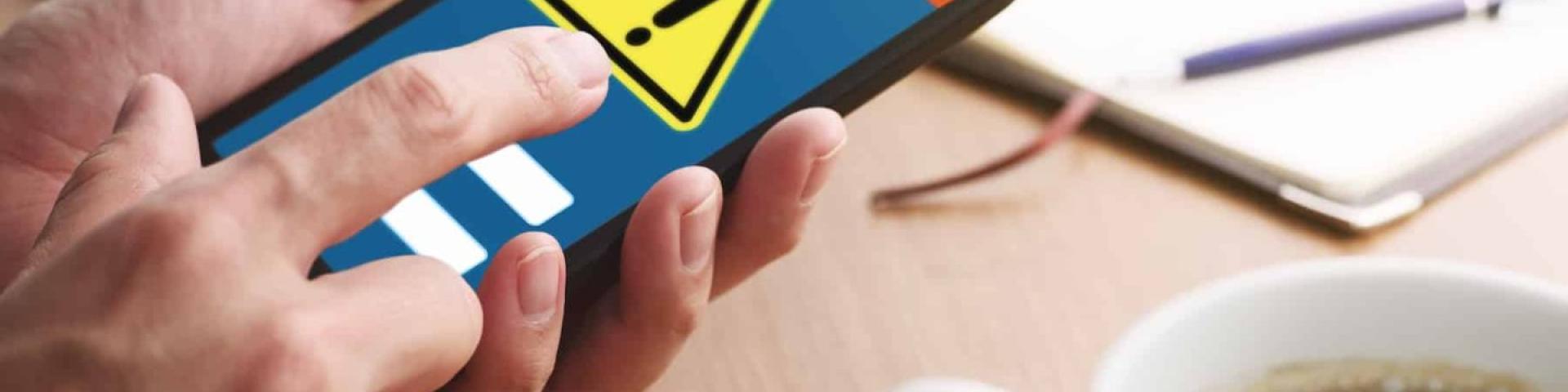 Person using smartphone with a warning icon; coffee and papers on a wooden table.