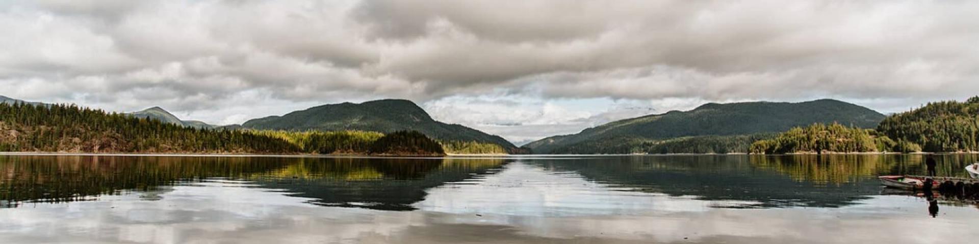 Calm lake with cloudy sky and distant forested hills reflecting on the water.