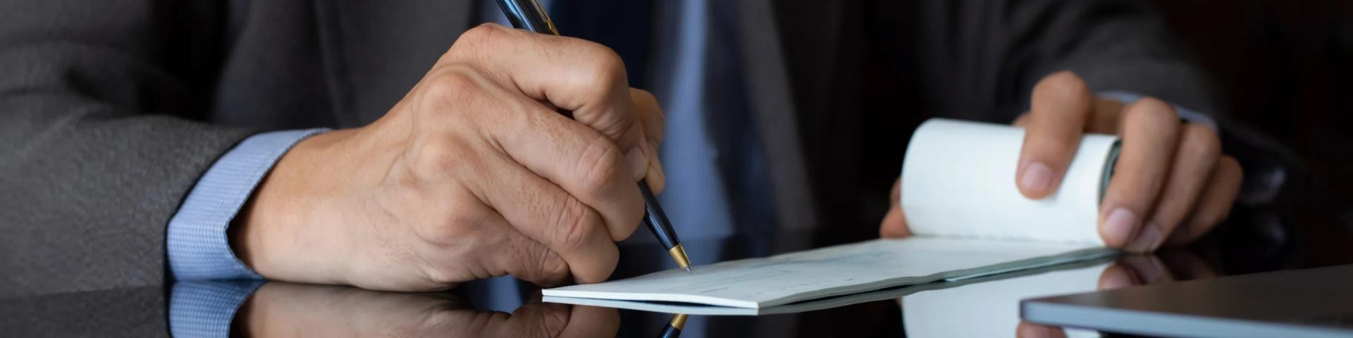 A person in a suit writes on a notepad while sitting at a reflective surface.