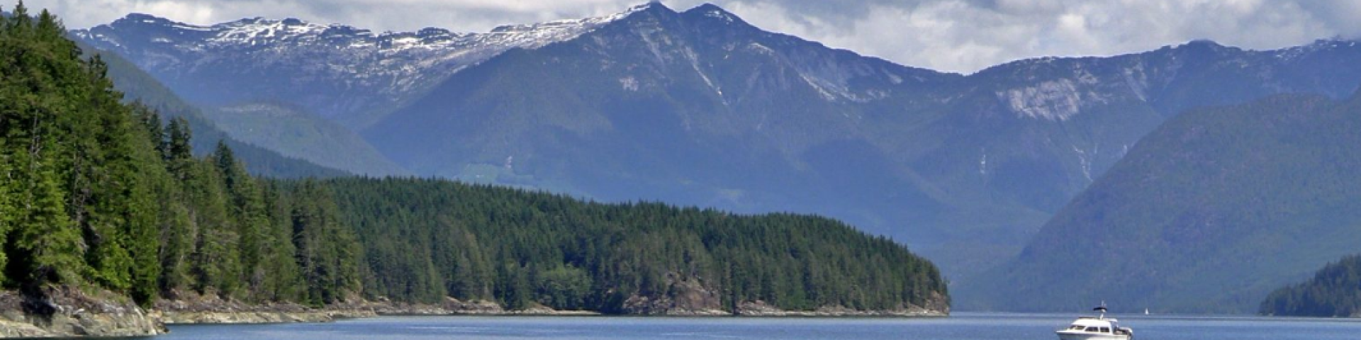 Calm water with a small boat, surrounded by lush green forests and distant mountains under a cloudy sky.