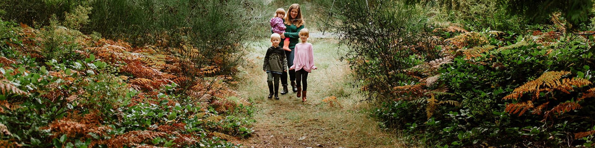 A woman and two children walk along a forest path, surrounded by greenery and autumn foliage.