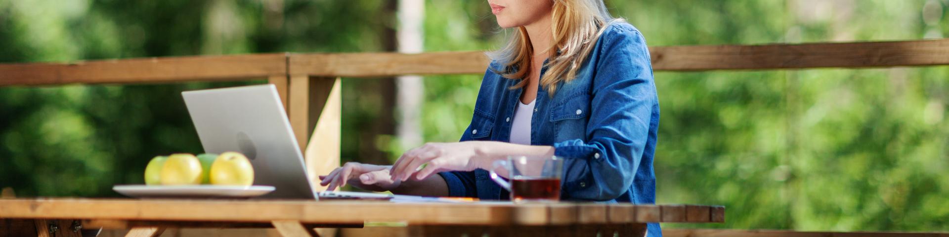 Woman working on a laptop at a wooden table surrounded by greenery.