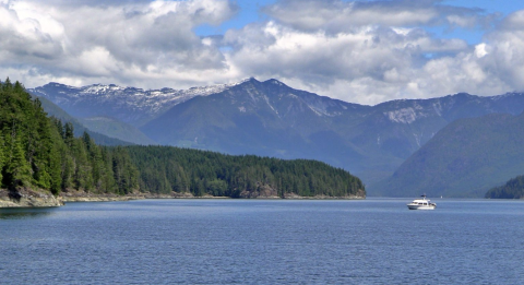 Calm water with a small boat, surrounded by lush green forests and distant mountains under a cloudy sky.
