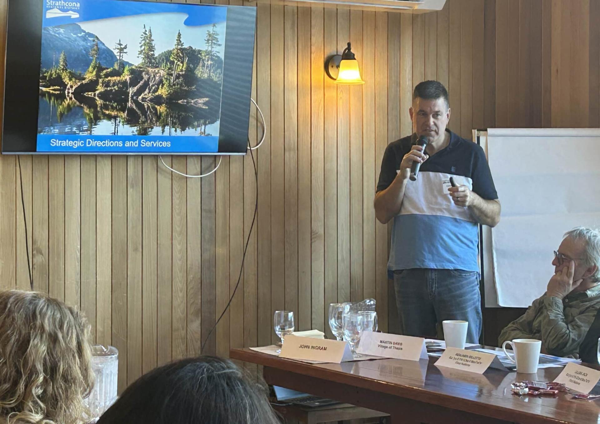 Man presenting with a microphone in a wooden-paneled room, screen displaying landscape image.