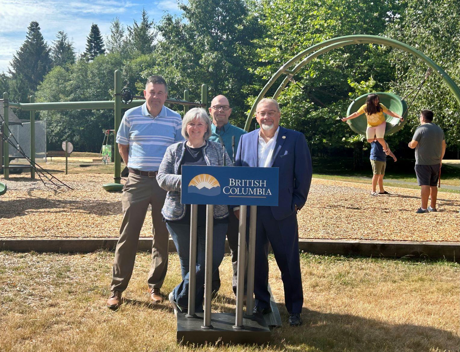 Four people stand at a podium outdoors near a playground with children playing in the background.