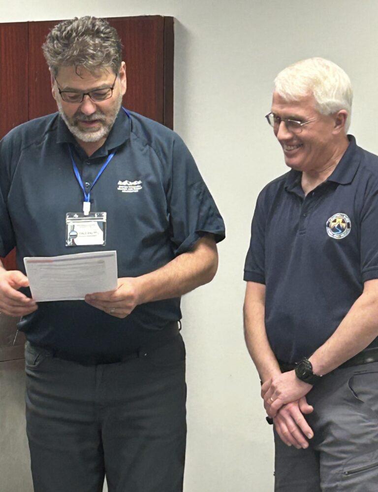 Two men in blue shirts, one reading a paper, both smiling.
